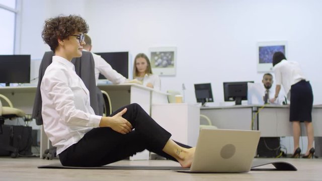 Wide shot of barefoot businesswoman with short curly hair sitting on yoga mat, stretching her body and using laptop while her colleagues working on background