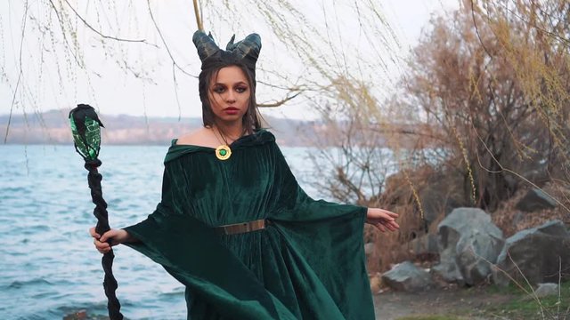 young cute girl with creative make-up poses for photographer by the lake, lady with big black horns on head in image of magical vampter in long flying emerald velor dress-mantle, wind chant and storm