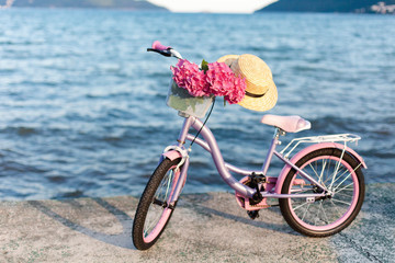 Fototapeta premium Female romantic bicycle with pink hydrangea flowers and straw hat on sea beach. Concept of children riding on coastline, tenderness, childhood, summer.