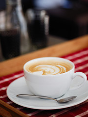 A big cup of freshly brewed cappuccino with latte art heart coffee on wooden table, red plaid tablecloth in cozy craft cafe with shallow depth of field and copy space.