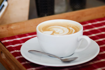 A big cup of freshly brewed cappuccino with latte art heart coffee on wooden table, red plaid tablecloth in cozy craft cafe with shallow depth of field and copy space.