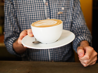 Close up of barista man hands serving freshly brewed cappucino coffee while standing in coffee shop. Focus on male hands holding a big white ceramic cup of coffee with copyspace