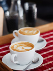 A two big cups of freshly brewed cappuccino with latte art heart coffee on wooden table, red plaid tablecloth in cozy craft cafe with shallow depth of field and copy space.