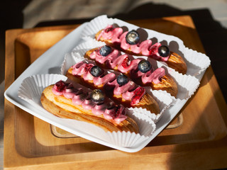 Craft delicious homemade eclairs sprinkled with berry cream, powdered sugar and chocolate drops on an square white ceramic plate on a wooden rustic old table, angle view from above. Cafe shop dessert
