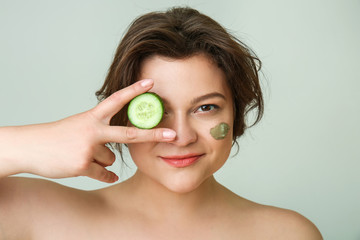 Portrait of beautiful plus size woman with cosmetic clay on her face and cucumber against light background