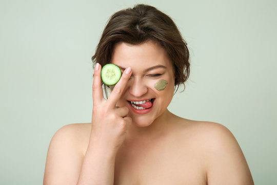 Portrait of happy plus size woman with cosmetic clay on her face and cucumber against light background - Powered by Adobe