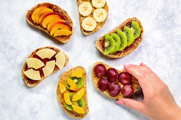 Woman hand putting breakfast toasts with peanut butter, strawberry jam, banana, grapes, peach, kiwi, pineapple, nuts