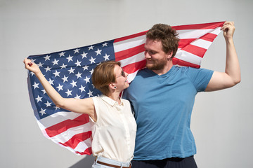 Young smiling caucasian couple of americans holding USA flag behind and looking at each other