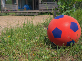 Close-up of a red soccer ball for children on the green grass in front of a country house. Porch of the house in the background. Space for text