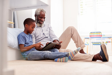 Grandfather Sitting With Grandson In Childs Bedroom Using Digital Tablet Together