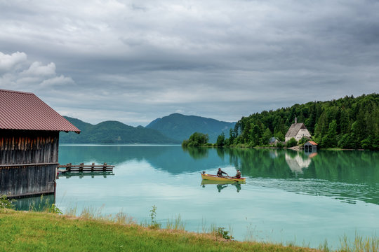 Der gr&uuml;n-t&uuml;rkise Walchensee in Bayern mit Ruderboot, Anglern, Bootshaus bei wolkigem Himmel