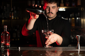 Professional male bartender pouring a transparent alcohol into the cocktail glass from the steel shaker