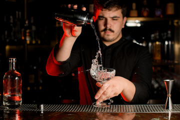 Professional bartender pouring a transparent alcohol into the cocktail glass from the shaker