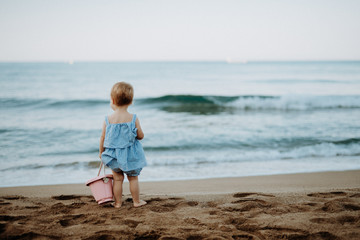 A rear view of small toddler girl standing on sand beach on summer holiday.