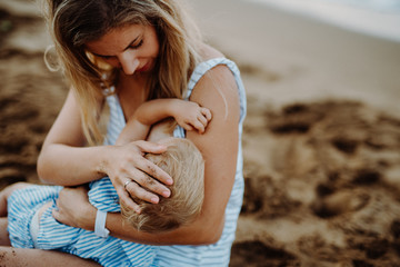 Young mother breasfeeding toddler daughter on beach on summer holiday.