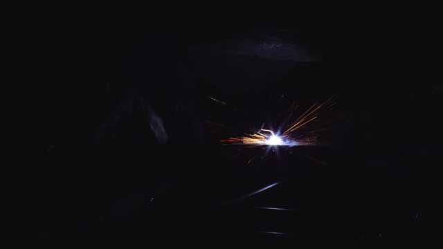 A blacksmith with a welding mask and welders leathers, is welding metal products in the garage, fire sparks fly to the sides. Dark mood. Slow Motion shot with Red Monstro 8K.