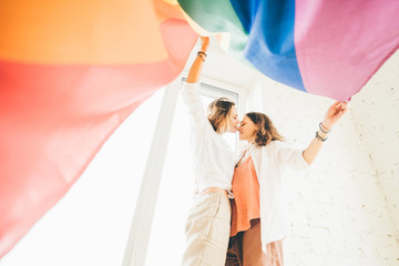 Beautiful female young lesbian couple in love standing near the window with the rainbow flag....