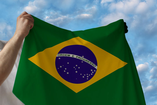 beautiful national flag of the country of Brazil in male hands against the blue sky with clouds