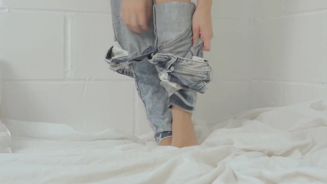 Closeup of woman's legs while taking off denim jeans trousers isolated on bed over white wall background
