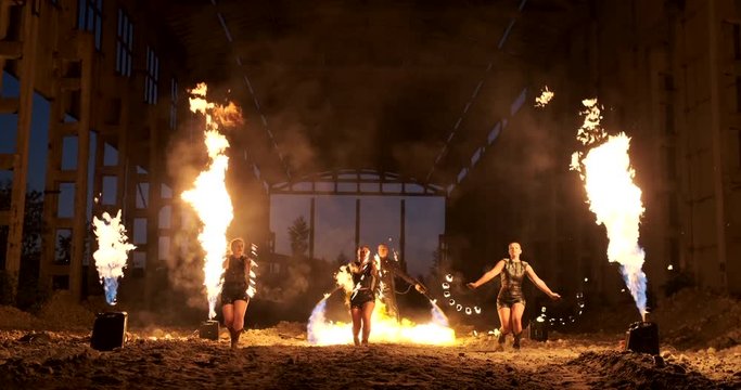 Four women in leather clothes with fire dance and show fire show a man with a flamethrower in the back plays with the flame in slow motion.