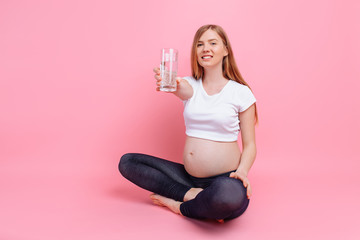 Pregnant girl holding a glass of water on the background of a pregnant belly, on a pink background