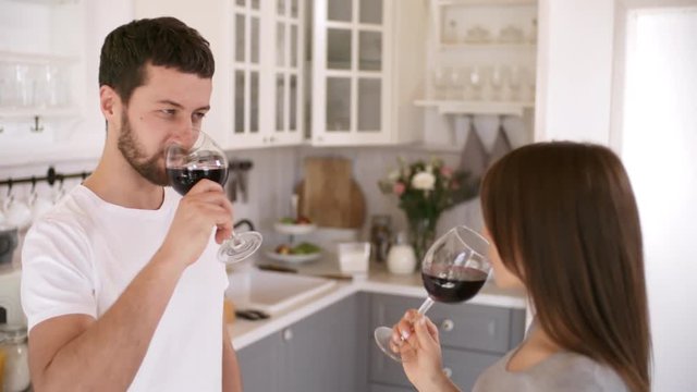 Handheld shot of loving couple standing in kitchen on lazy morning and clinking glasses of red wine, then drinking and kissing