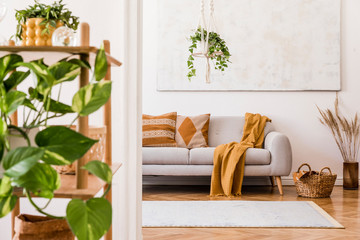 The stylish boho interior of living room in nice apartment with gray sofa, honey yellow plaid and pillows, a lot of plants and elegant accessories. Handmade macrame shelf planter hanger and paintings.