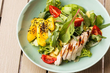 Salad with grilled chicken, mango, lettuce, avocado, tomatoes, arugula, cheese sause on a white plate on wooden background