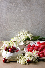 Fresh cherry and raspberry berries in ceramic mug and plate, elderflowers, jug of cream on kitchen table with gray wall at background. Copy space