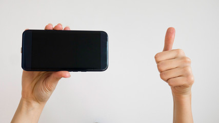 Caucasian female holds a black mobile phone with right hand and gives thumbs up with left hand on a white background  