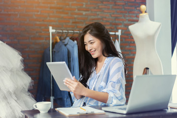 Fashion designer drawing new sketches collection and sent a business e-mail to client. Beautiful young Asian woman working using laptop and digital tablets in Designers Stylish Showroom the workshop