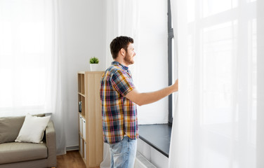people concept - young man opening window curtain at home