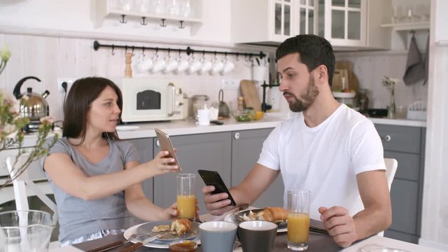 Handheld shot of young woman and bearded man sitting at kitchen table and showing each other funny pictures on mobile phones while having breakfast together