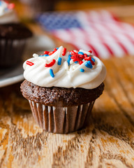 Close up of a mini chocolate cupcake with red, white, and blue sprinkles.  Additional cupcakes and US flag blurred in the background.  Wooden table.
