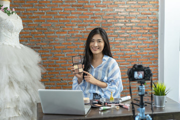Young Asian cute artist woman beauty vlogger or blogger doing a cosmetic makeup tutorial vlog with brushes looking camera, recording viral clip to share on social media live streaming for Internet