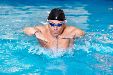 Fototapeta na wymiar Muscular swimmer young man in black cap in swimming pool, performing butterfly stroke .