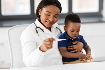 medicine, healtcare, pediatry and people concept - happy african american female doctor or pediatrician with thermometer measuring baby's temperature on medical exam at clinic
