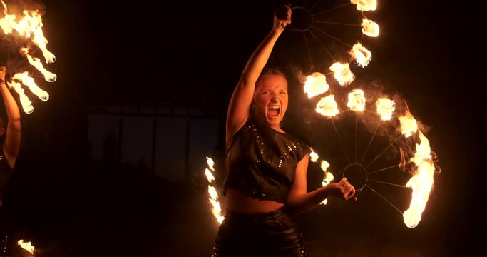 Three women with burning hoops dance with fiery torches in leather clothes in a dark hangar demonstrating a circus fire show in slow motion.