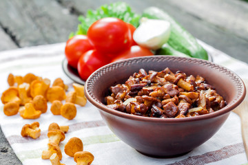 Fried chanterelles with onion in rustic bowl and plate with fresh vegetables for salad on background