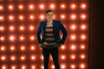 Fototapeta na wymiar Young male standing against illuminated wall. Trendy man standing with hands on hips and looking at camera on red background with light bulbs