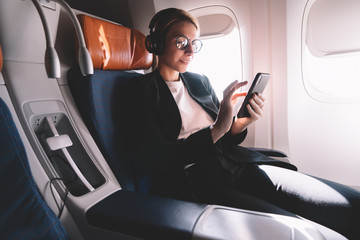 Young caucasian woman sitting near window with smartphone connected to wifi internet during flight on board, female passenger in headphones for noise cancellation search songs in online shop on app © BullRun