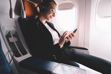Hipster girl flight passenger connected by smartphone to wireless network inside airplane cabin, smiling female in bluetooth headphones for noise cancellation enjoying music songs on mobile phone app - obrazy, fototapety, plakaty