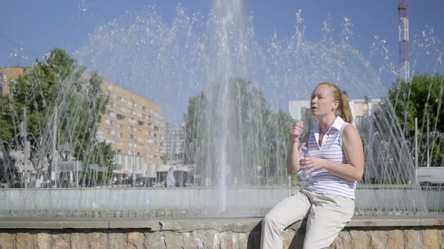 girl, exhausted from stuffiness, waves her hand near city fountain. woman sits by fountain on sunny day and suffers from heat.