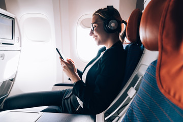 Cheerful female passenger in headphones for noise cancellation watching online movie during...