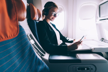 Happy smiling woman passenger in headphones for noise cancellation enjoying intercontinental flight in first class with high speed wifi connection on board using touch pad and browsing internet