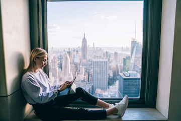 Trendy dressed hipster girl using digital tablet while sitting on hotel window sill with breathtaking scenery view of New York cityscape. Female digital nomad working remote on freelance, millennials