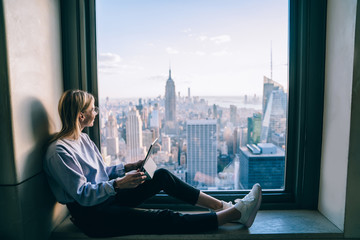 Happy smiling female tourist enjoying scenic views from hotel window of famous New York landmark during her vacation holidays. Attractive hipster girl sitting with digital tablet in coworking office