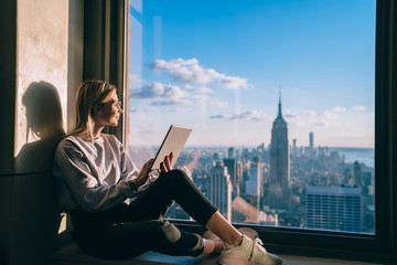 Smiling casual dressed coworking visitor working on freelance with touch pad and enjoying beautiful view of New York city. Caucasian hipster girl enjoying skyline view during vacation holidays in USA