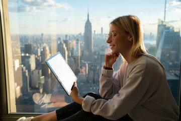 Smiling female traveler reading interesting electronic book on tablet computer while sitting on window sill with panoramic views on New York city. Hipster girl browsing social network on touch pad