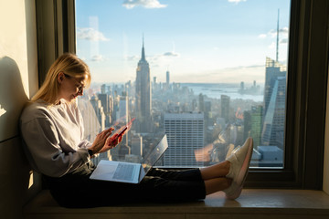 Trendy caucasian female texting message on smartphone while sitting on office window sill with...
